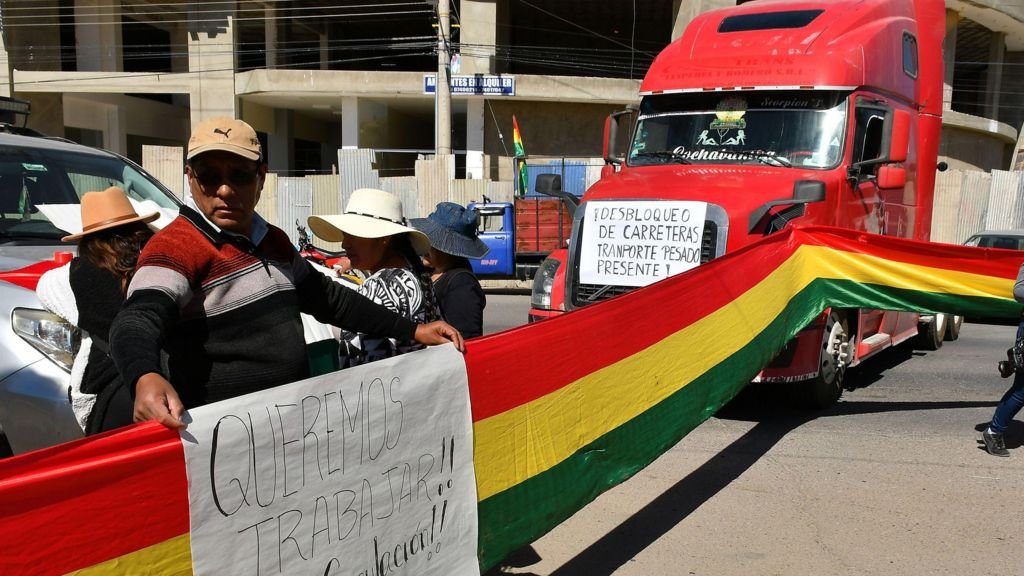 Protesters hold a flag and sign next to a road block with several vehicles in Bolivia