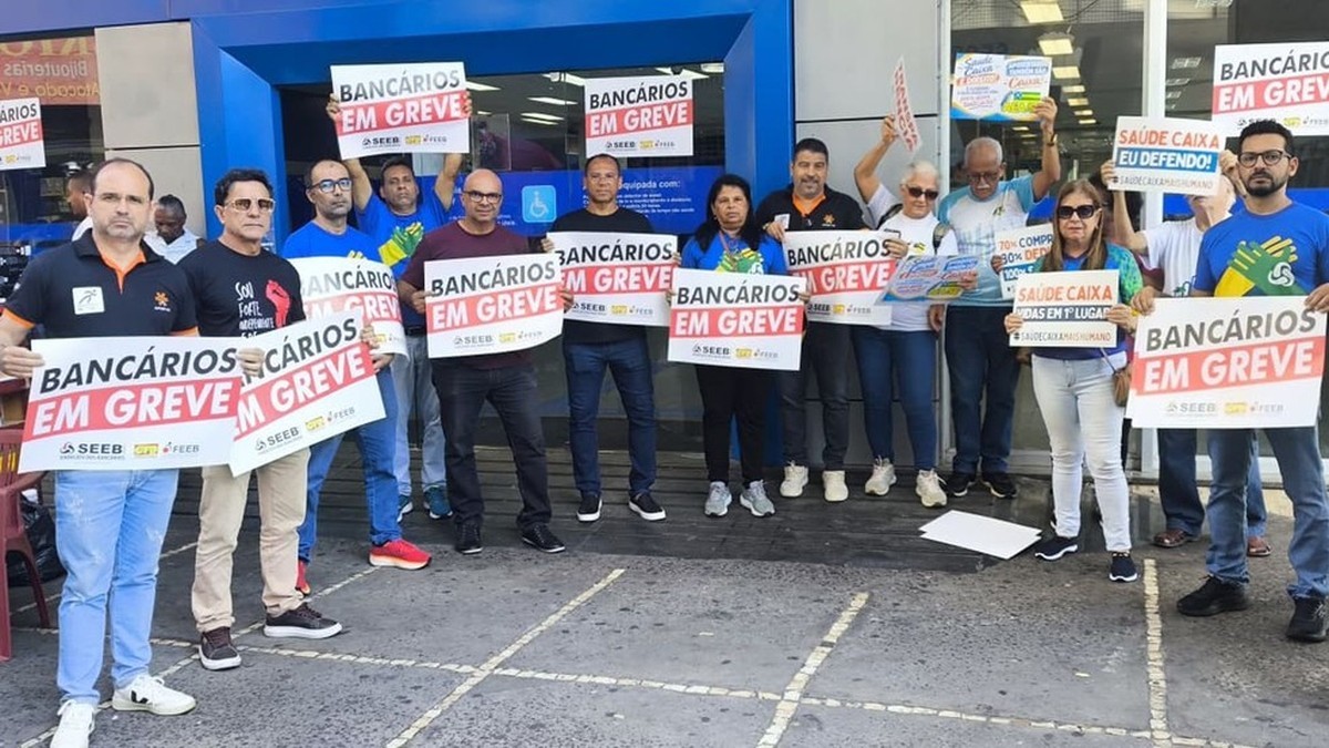 Bank workers on strike hold printed banners and signs outside a bank branch entrance. The banners read Bancarios em Greve and Saude Caixa e Defender o SUS. Photo: Personal archive via G1